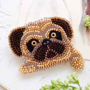 Può includere: Spilla a forma di carlino fatta a mano con perline marroni, beige e nere. Il carlino è raffigurato sdraiato. Sullo sfondo è visibile un'illustrazione ad acquerello di cani. Accessorio unico.