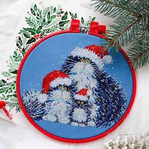 May include: A cross-stitch embroidery project in a red circular hoop featuring three fluffy cats with yellow eyes wearing red and white Santa hats against a blue background. The cats are depicted with detailed stitching in shades of grey and black.