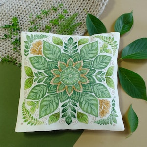 May include: A square white throw pillow featuring a detailed cross-stitch embroidery of a symmetrical botanical mandala. The design showcases various shades of green and gold leaves, ferns, and foliage arranged in a circular, radiating pattern.