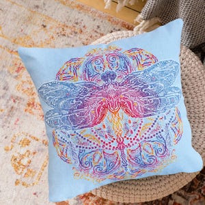Op de afbeelding: Een lichtblauw vierkant kussen met een gedetailleerd libellenontwerp. De libel is weergegeven in tinten blauw, paars, roze en geel. Het kussen ligt op een geweven oppervlak, geschikt voor interieurdecoratie.