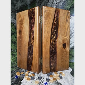 Puede incluir: Juego de backgammon de madera con un acabado de vetas naturales. El tablero presenta una incrustación oscura en forma de río. Incluye fichas de juego en tonos neutros y dos dados azules. El tablero de backgammon está abierto, mostrando la superficie de juego.