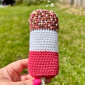 May include: A handmade crochet ice pop in shades of brown, white, and red. The top section is brown with colorful sprinkles, followed by a white middle section, and a red base. A beige stick extends from the bottom.