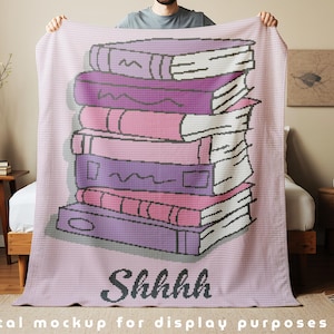 Puede incluir: Una manta rosa claro con un diseño de libros apilados en tonos morados y rosas. La palabra "Shhhh" está impresa en negro. La manta se muestra en una cama.