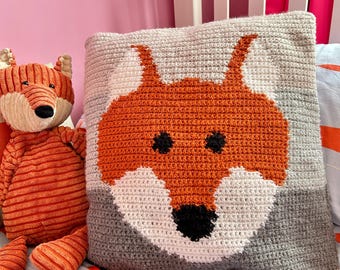 Könnte beinhalten: Ein gehäkeltes quadratisches Kissen mit einem Fuchsmotiv in Orange, Weiß, Schwarz und Grau. Daneben befindet sich ein Stofffuchs-Spielzeug mit orangefarbenen und cremefarbenen Streifen. Der Hintergrund umfasst eine rosa Wand und einen weißen Bettrahmen.