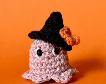 May include: A handmade crochet octopus in lavender with a black witch hat and orange accents. The octopus has two black eyes and a small, rounded body. The background is a solid orange.