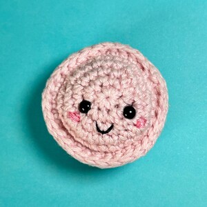 May include: A handmade, light pink crocheted circle with a smiling face. The face features black eyes, pink cheeks, and a curved black mouth. The background is a solid turquoise colour.
