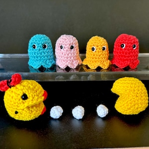 May include: Crocheted Pac-Man characters and ghosts in vibrant colors. The ghosts are in blue, pink, yellow, and red, each with black eyes. Pac-Man and a female Pac-Man are yellow, with white pellets. A fun, handmade item.
