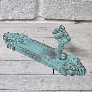May include: A distressed, turquoise-colored key holder with a vintage aesthetic. The key holder features a rectangular base with ornate detailing and a key-shaped hook. The piece has a weathered appearance, suggesting an antique or rustic style.