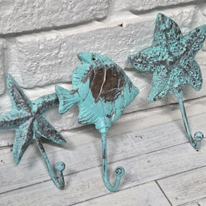 May include: Three decorative wall hooks in the shape of a starfish and a fish. The hooks are made of metal with a distressed turquoise finish and brown accents. The hooks are mounted on a white brick wall.