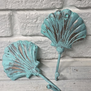 May include: Two decorative seashell-shaped wall hooks. The hooks are painted in a distressed turquoise finish with bronze accents. The hooks are made of metal and are designed to be mounted on a wall for hanging items.