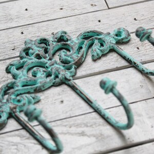 May include: A wall-mounted metal hook rack featuring two curved hooks and an ornate, scrollwork backplate. The metal has a distressed finish with a bright turquoise patina layered over dark brown, showing visible wear and texture throughout.