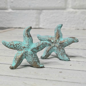 May include: Two decorative starfish figurines with a distressed turquoise and gold finish. These nautical-themed ornaments are made of a solid material and are displayed on a light wood surface. The starfish have five points and are slightly raised.