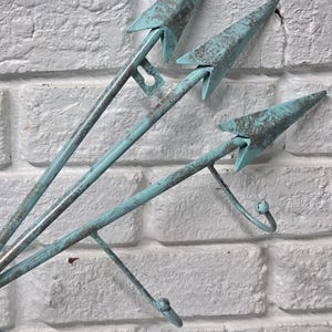 May include: A wall-mounted metal arrow-shaped decor piece. The arrows are painted in a distressed turquoise color with bronze accents. The design includes hooks for hanging items. The background is a white brick wall.