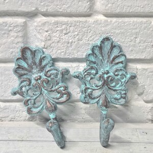 May include: Two ornate wall hooks with a distressed turquoise and bronze finish. The hooks feature a detailed, scrolling design and a curved hook at the bottom. The hooks are made of metal and are suitable for hanging items.