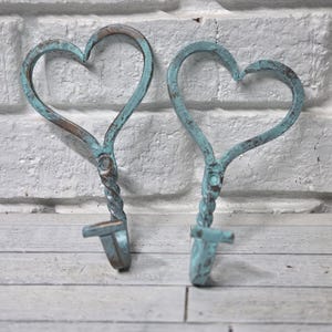 May include: Two heart-shaped wall hooks with a distressed teal finish. The hooks have a twisted stem and a small hook at the bottom. The background is a white brick wall and a wooden surface.