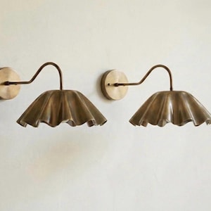 May include: Two wall-mounted sconces with a vintage brass finish. Each sconce features a circular base, a curved arm, and a ruffled, bell-shaped shade. The fixtures are designed to provide ambient lighting and add a touch of elegance to any space.