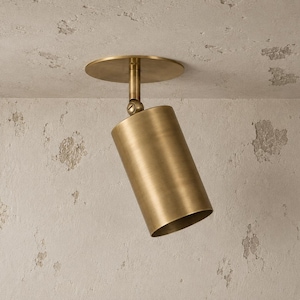 May include: A cylindrical brass spotlight fixture featuring a circular mounting plate and an adjustable swivel joint. The metal surface has a brushed finish, and the light is oriented at an angle, extending from a flat, textured ceiling surface.