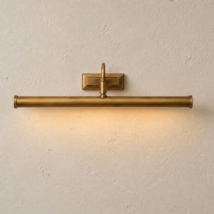 May include: A horizontal wall-mounted picture light with an antique brass finish. The fixture features a rectangular backplate, a central vertical arm, and a long cylindrical tube that emits a warm, downward-facing glow across the wall surface.
