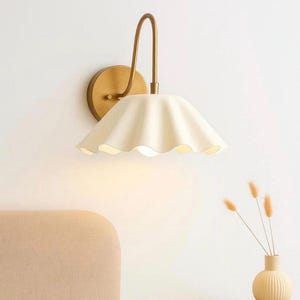 May include: A wall-mounted lamp featuring a curved brass-toned metal arm and a circular wall plate. The light fixture includes a wide, off-white, scalloped-edge shade that directs light downward, creating a soft glow against the wall surface.