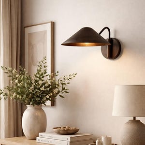 Puede incluir: Un aplique de pared de color bronce con una pantalla cónica y un brazo curvo está montado en una pared de color claro. La escena incluye un jarrón con flores, una lámpara de mesa y objetos decorativos.