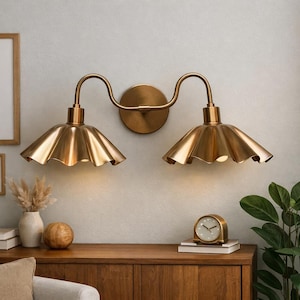 Può includere: Applique da parete in ottone con due luci, ciascuna con un paralume a balze. L'apparecchio è montato su una parete colorata chiara sopra un mobile in legno. L'applique aggiunge un tocco di eleganza vintage.