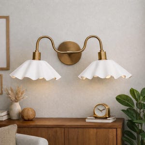 May include: A double-light wall sconce with a brass finish and two white, scalloped-edge shades. The sconce is mounted on a light-colored wall above a wooden cabinet. Decorative items like a vase, clock, and artwork are also visible.