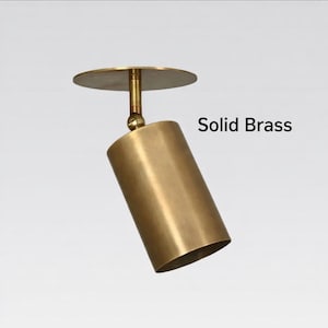 May include: A solid brass ceiling spotlight. The fixture features a cylindrical light head, a round mounting plate, and a connecting arm. The text "Solid Brass" is visible.