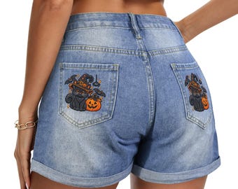 May include: Blue denim shorts featuring a high-waisted design with rolled cuffs. The back pockets are decorated with embroidered patches of a black cat wearing a witch hat, an orange jack-o'-lantern, and small bats on a dark background.