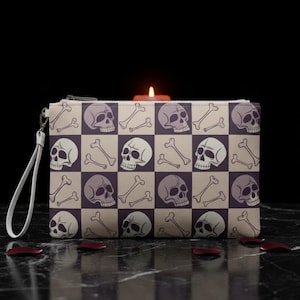 May include: A rectangular clutch bag with a zip and wrist strap. The bag displays a repeating pattern of skulls and crossbones on a checkered beige and purple background. A lit candle is positioned behind the clutch.