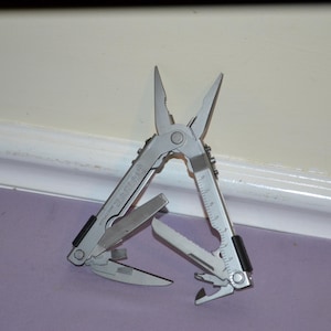 Pode incluir: Uma multi-ferramenta prateada com alicate, faca, serra e outras ferramentas. A ferramenta está parcialmente aberta, exibindo suas várias funções. A ferramenta está em uma superfície roxa.