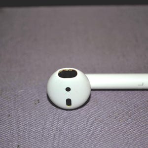 May include: A white wireless earbud with a rounded head and a stem. The earbud has a small black dot and a rectangular opening on the front. The stem has the letter "L" printed on it.