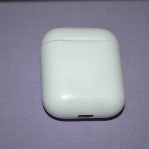 Puede incluir: Un estuche de carga blanco y rectangular para auriculares inalámbricos. El estuche es brillante y tiene un pequeño puerto de carga en la parte inferior. El estuche está cerrado y descansa sobre una superficie púrpura.
