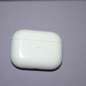 May include: A white, glossy, rectangular charging case for wireless earbuds. The case is closed and has rounded corners. It sits on a light purple surface.