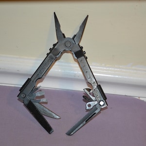 Könnte beinhalten: Ein silbernes und schwarzes Multitool mit Zange, Messer und anderen Werkzeugen. Auf einer Seite ist ein Lineal eingraviert. Die Zange ist geöffnet, andere Werkzeuge sind ausgeklappt. Für verschiedene Aufgaben konzipiert.
