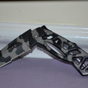 May include: A folding knife with a camouflage pattern on the blade and handle. The blade has a serrated edge and the word "BUCK" is printed on the blade. The handle has a skeletal design.