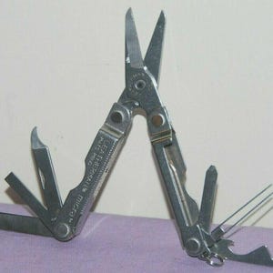 Puede incluir: Una multiherramienta Leatherman Micra plateada con múltiples herramientas extendidas. Las herramientas incluyen tijeras, un cuchillo, una lima de uñas, un destornillador, pinzas y un abrebotellas. La multiherramienta está abierta y se muestra sobre un fondo morado y blanco.