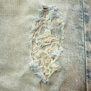 May include: Close-up of light blue denim fabric with a distressed area patched with white lace. The lace patch is oval-shaped and adds a delicate contrast to the rugged denim texture. The edges of the patch are frayed.