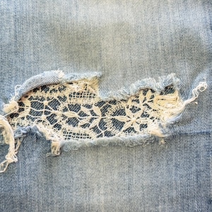 May include: Close-up of light blue denim jeans with a distressed area. The distressed area is patched with white floral lace, creating a unique design. Frayed edges and a textured appearance add to the jeans' aesthetic.