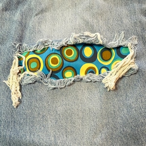 May include: Close-up of light blue denim fabric with a distressed patch. The patch features a vibrant blue fabric with a retro pattern of yellow, brown, and green circles. The edges of the denim are frayed, and the patch is secured with white thread.