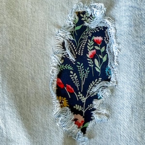 May include: Close-up of a light blue denim fabric with a tear, patched with a floral fabric. The patch features a dark blue background with red, pink, and white flowers and green leaves. The edges of the patch are frayed.
