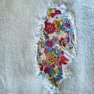 May include: Close-up of light blue denim fabric with a distressed patch. The patch is a floral print with pink, yellow, and blue flowers. The edges of the denim are frayed, adding a vintage look.