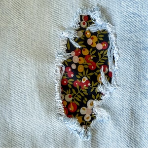 May include: Close-up of light blue denim fabric with a tear, patched with a floral fabric. The patch features a dark background with red, yellow, and white flower and leaf patterns. The edges of the denim are frayed.