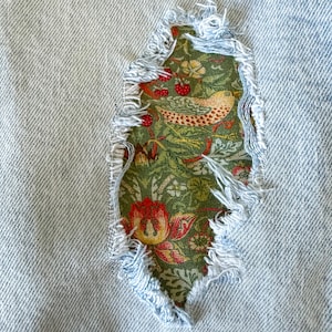 May include: Close-up of light blue denim fabric with a distressed, torn patch. The patch reveals a green floral fabric with a bird and red berries. The edges of the denim are frayed, adding a vintage, worn look.