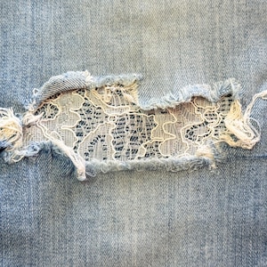 May include: Close-up of light blue denim jeans with a distressed area. The distressed area is patched with white lace, creating a unique design. The edges of the denim are frayed, adding to the worn look.