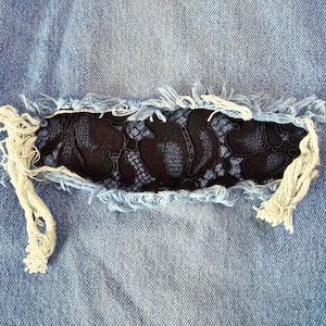 May include: Close-up of light blue denim fabric with a distressed patch. The patch features a black floral lace design, framed by frayed denim edges and white threads. The overall aesthetic is a blend of casual and decorative.