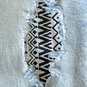 May include: Close-up of distressed light blue denim jeans with a decorative patch. The patch features a black and white geometric pattern, visible through the ripped fabric. Frayed edges add texture to the design.