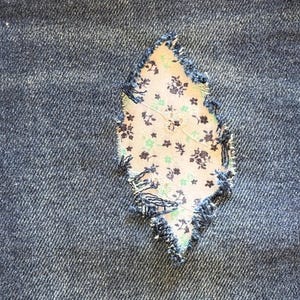 May include: Close-up of a denim fabric with a diamond-shaped patch. The patch is made of a floral patterned fabric in beige, green, and brown. The edges of the patch are frayed, and the denim is a dark blue color.