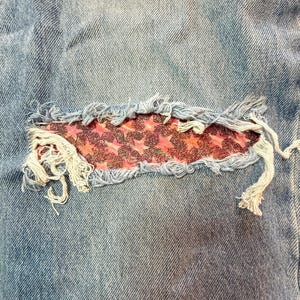 May include: Close-up of light blue denim jeans with a distressed patch. The patch is a rectangular shape with a pink and red star pattern. Frayed edges and loose threads add to the worn aesthetic.