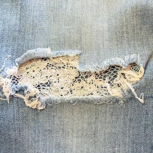 May include: Close-up of light blue denim jeans with a distressed knee. The tear reveals a delicate white lace patch underneath, adding a contrasting texture and design element. Frayed edges surround the tear.