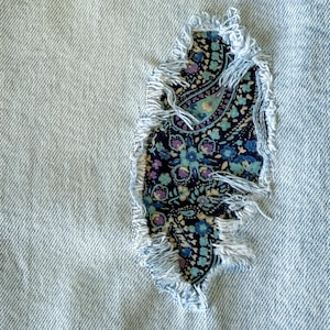 May include: Close-up of light blue denim fabric with a tear, revealing a paisley patch. The patch features a floral design in shades of blue, purple, and green. The edges of the tear are frayed.